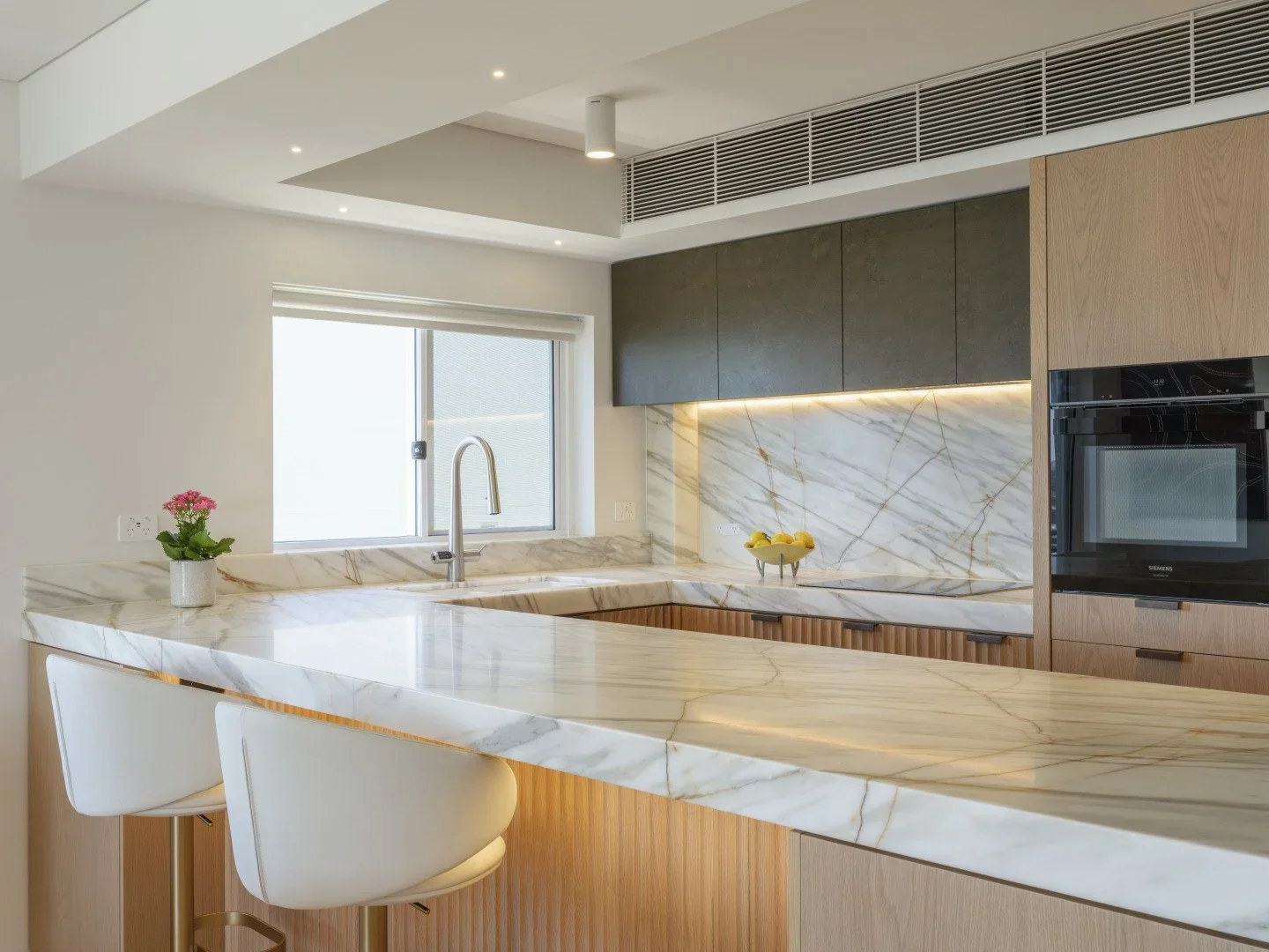 Modern kitchen with marble countertops, a window behind the sink, gray and wooden cabinetry, a built-in oven, and a small potted plant on the counter.