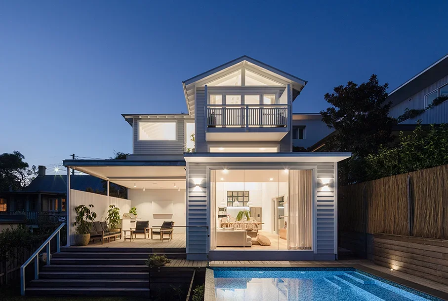 A modern two-story house with a balcony, large sliding glass doors, an outdoor seating area, a wooden privacy fence, and a swimming pool at dusk.