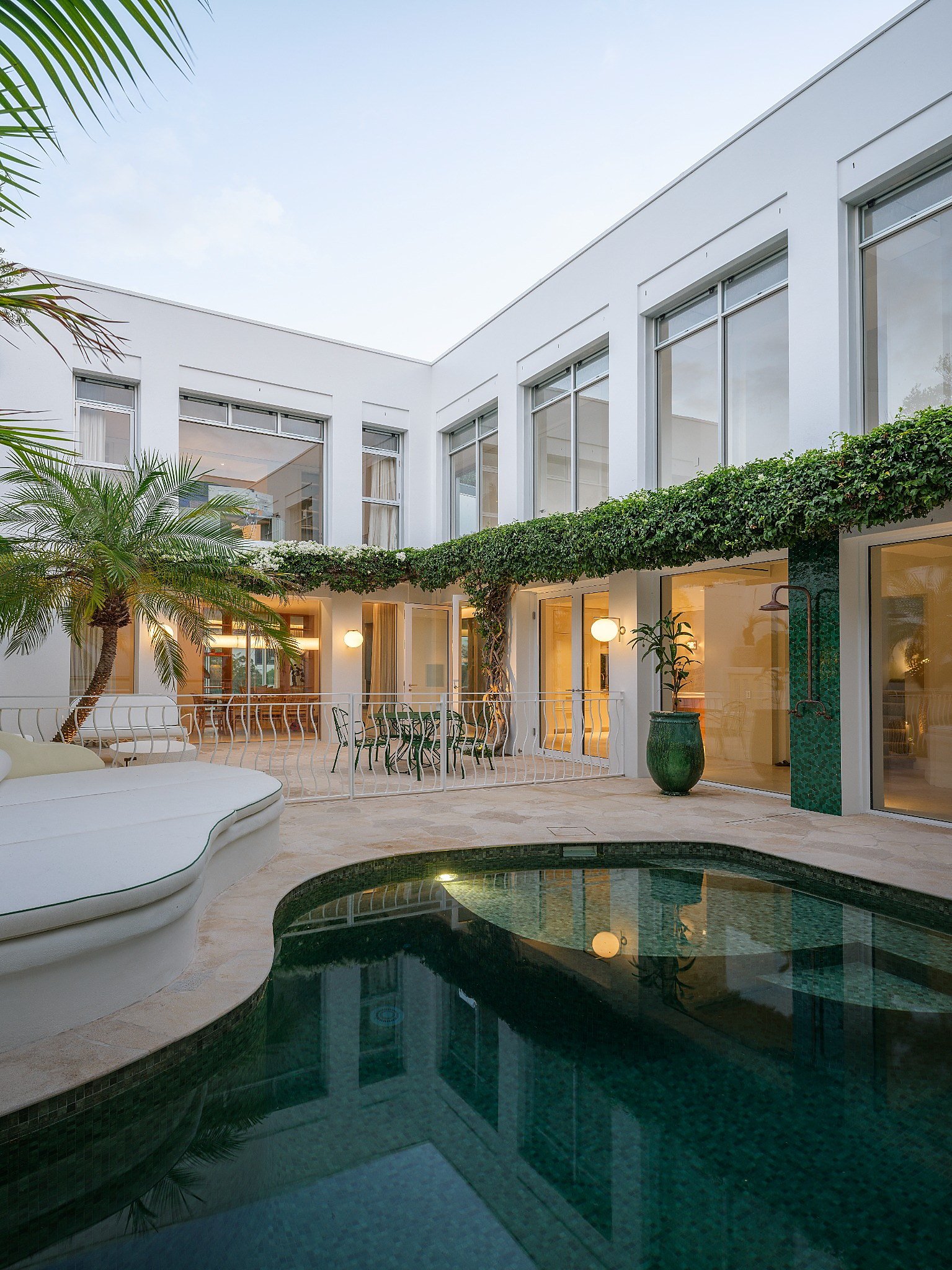A modern white residential building with large windows, outdoor seating area, a swimming pool, lush green plants, and string lights, during dusk.