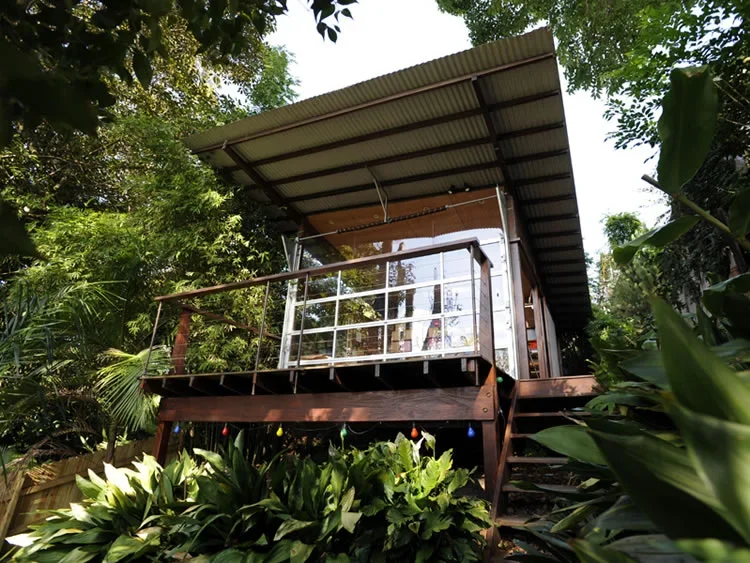 A small elevated house or cabin surrounded by lush green tropical plants, with a metal roof and a glass-paneled porch with metal railing, accessed by wooden stairs.