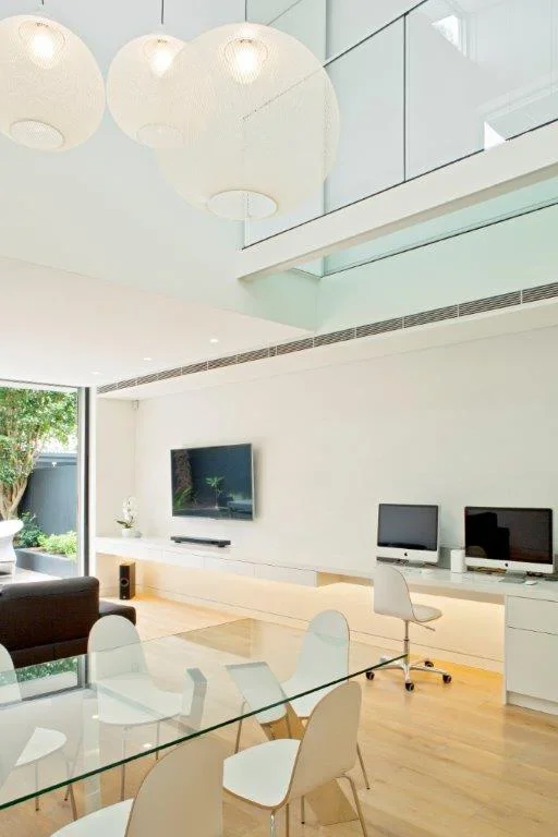 Modern living room with white walls, glass dining table with white chairs, a large TV mounted on the wall, and two desktop computers on a white desk. Large hanging pendant lights above.