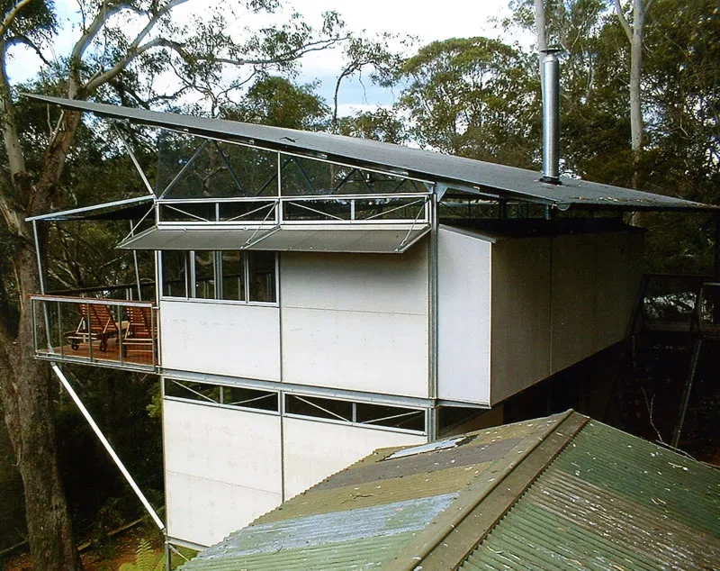 Modern house built on stilts with metal and glass balcony, surrounded by trees, with a sloped metal roof in an outdoor setting.