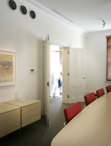 A dining room with a white wall, a closed scroll door, and a round table with red chairs. There is a window with brown blinds and some abstract artwork on the wall. A doorway leads outside, with a person partially visible.