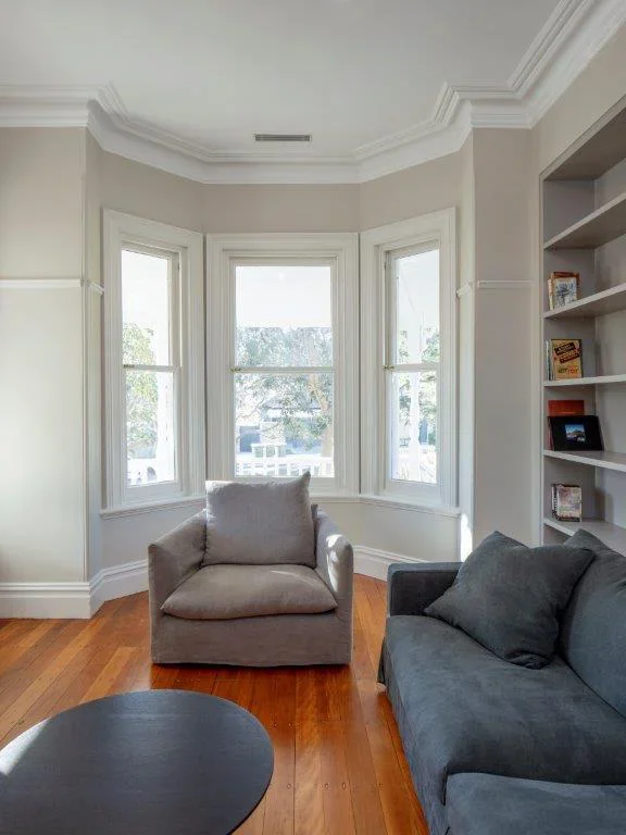 Living room with a large bay window, a beige armchair, a dark gray sofa with a pillow, a black coffee table, hardwood floors, white walls, and built-in bookshelves with books.