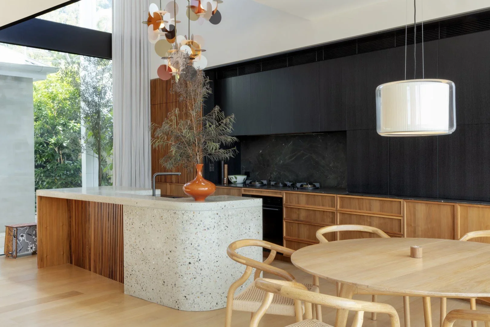 Modern kitchen with black cabinets, a terrazzo island, wooden furniture, hanging pendant lights, and large windows with greenery outside.