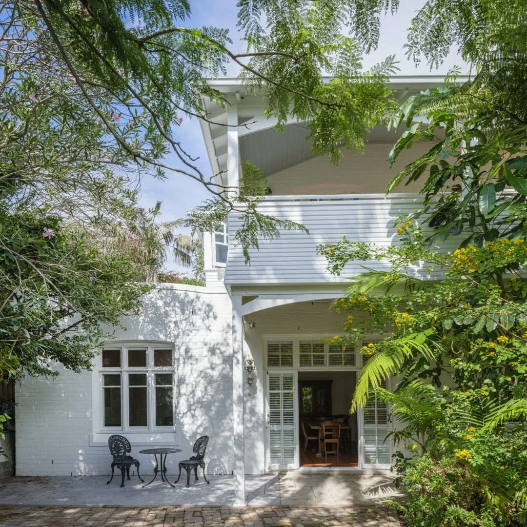 White two-story house with a patio area surrounded by lush green trees and plants, featuring a small outdoor table and two chairs, and open French doors leading into the interior.
