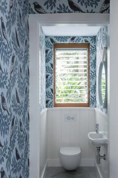 Small bathroom with bird and leaf-patterned wallpaper, a window with blinds, a toilet, a small sink, and a round mirror.