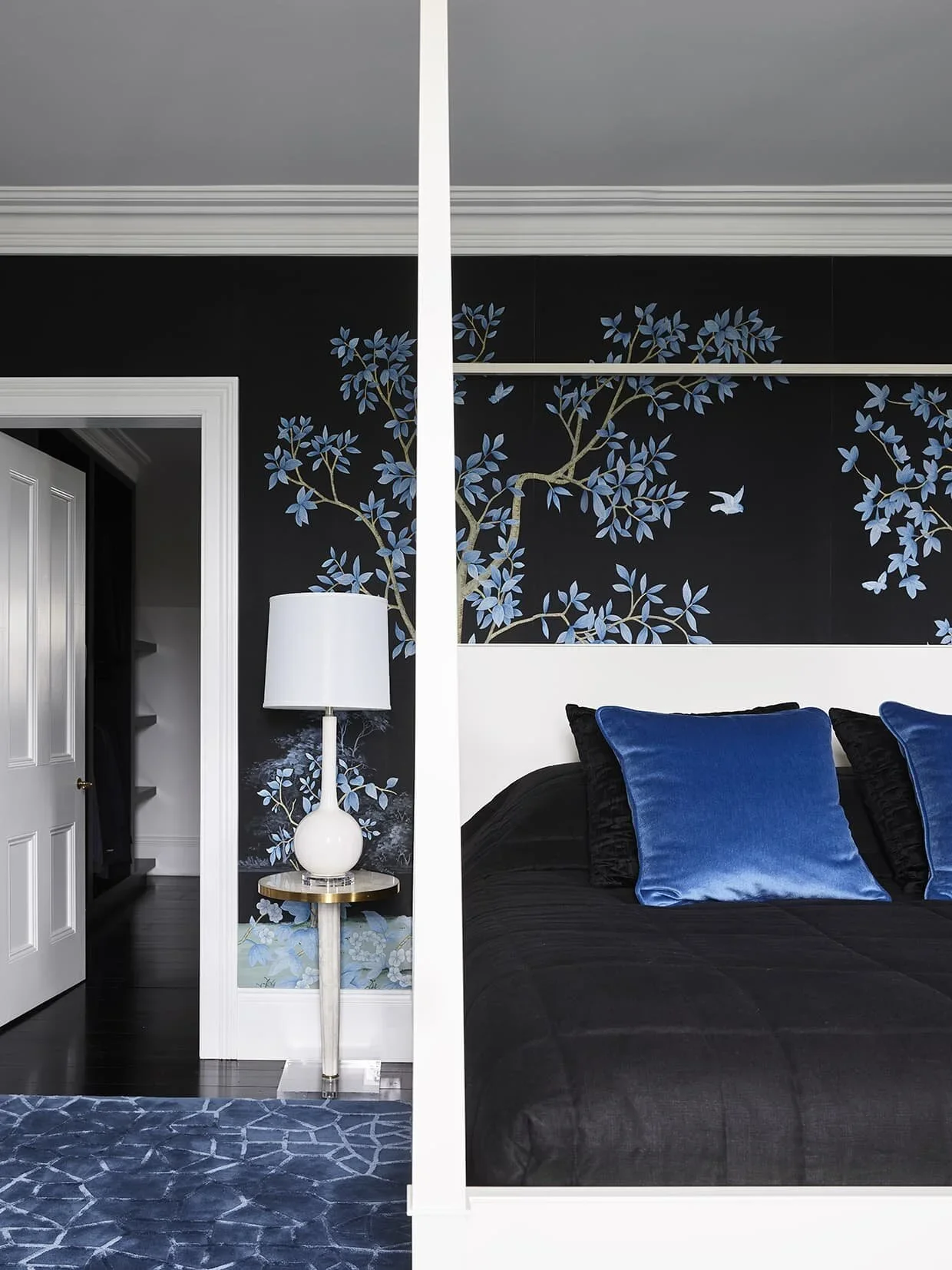 A bedroom with a dark wall featuring a blue and gray tree mural, a white door, a white bedside table with a white lamp, and a bed with black bedding and blue pillows. A blue patterned rug covers the floor.