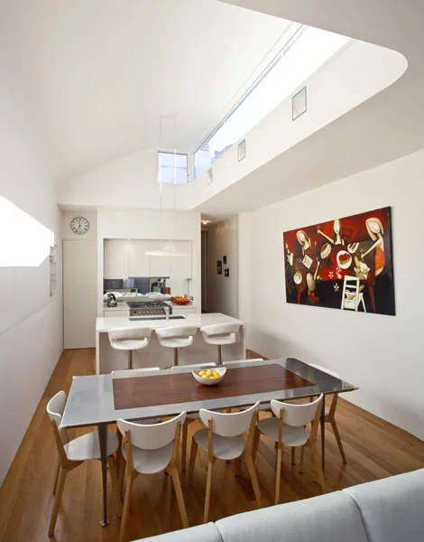Modern dining area with white chairs around a dark wooden table, kitchen in the background, colorful abstract painting on the wall, and natural light flooding in through a skylight.