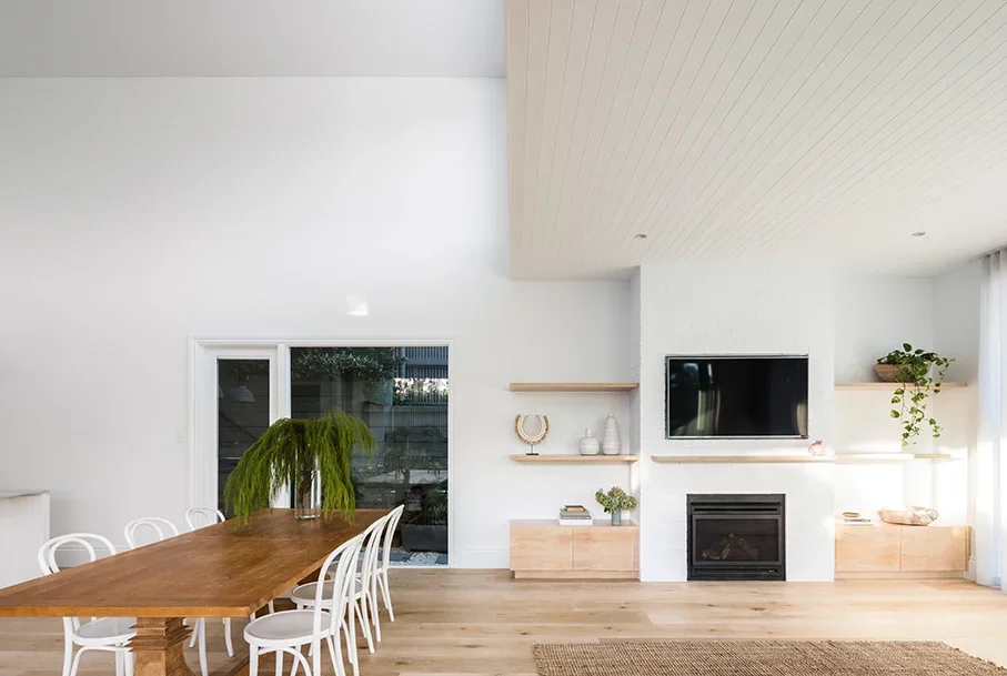 Modern living room with a wall-mounted TV above a fireplace, open shelves with decorative items, a potted plant, and a large wooden dining table with white chairs.