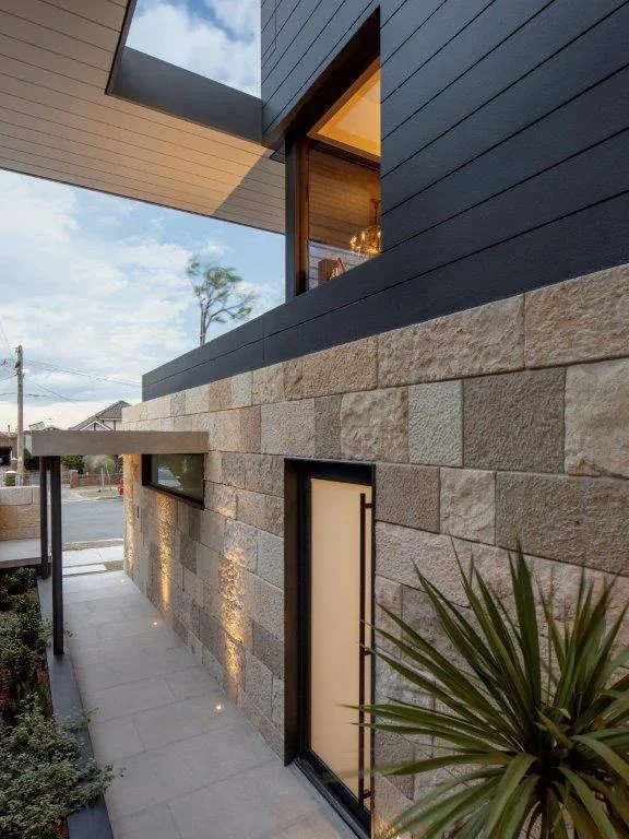 Modern building with beige stone exterior, black-framed glass door, narrow horizontal window, and dark siding, with a plant in the foreground.