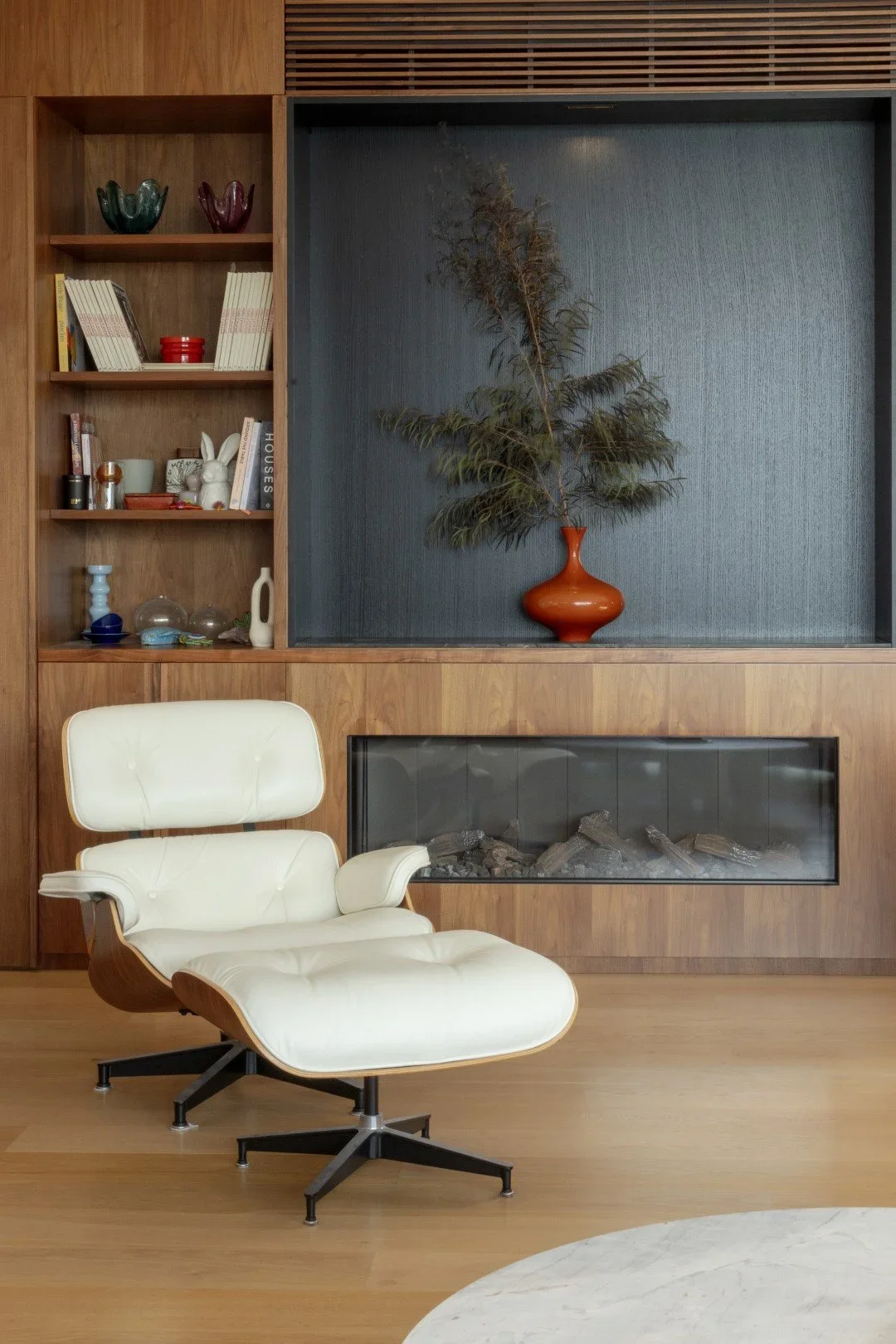 Modern living room with white Eames lounge chair, built-in wooden shelving, decorative items, fireplace, and an orange vase with green foliage.
