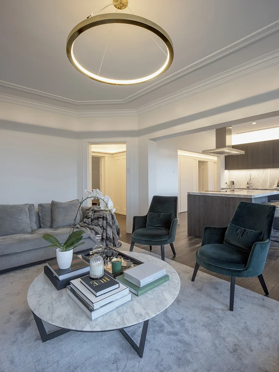 Living room with a grey sofa, two dark green armchairs, a round marble coffee table with books and decor, and modern lighting fixtures.