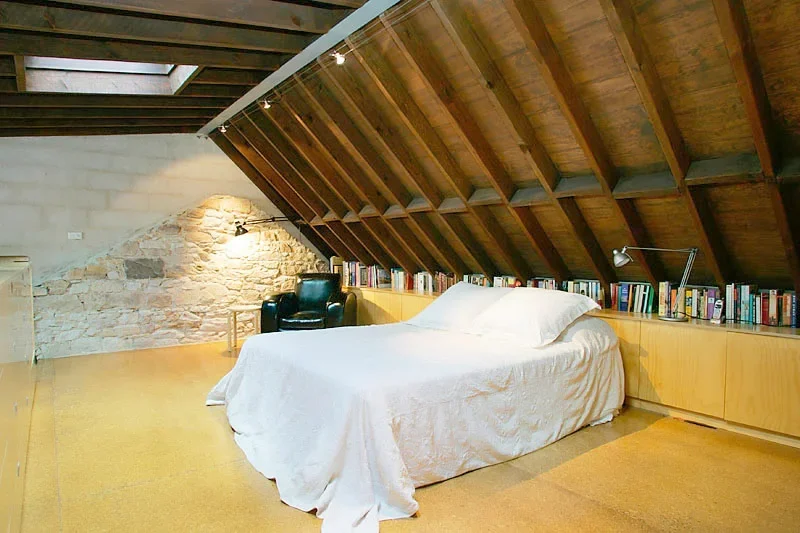 A cozy attic bedroom with a slanted wooden ceiling, a white bed with white linens, a bookshelf along the wall, a black armchair, and a stone accent wall.