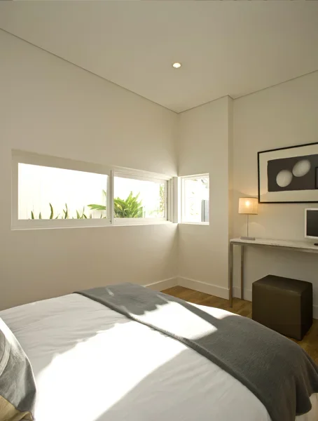 A bedroom with a bed, a window with a view of greenery, a desk with a computer, a framed black and white art piece, a lamp, and a small storage ottoman.