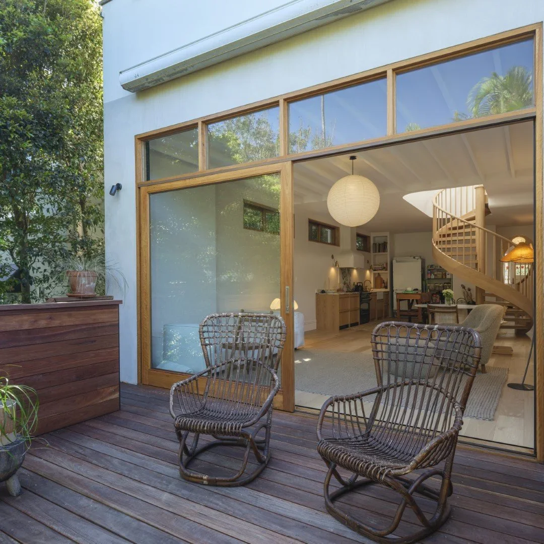 View of a wooden deck with two wicker rocking chairs outside a house with large glass sliding doors, revealing a cozy living room with a spiral staircase, dining area, and kitchen inside.
