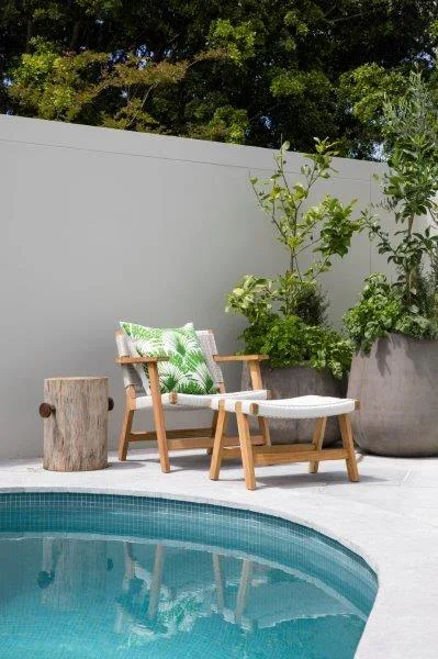 Poolside patio with a wooden lounge chair, a matching footstool, a decorative pillow with green leaf pattern, a small wooden side table, and large potted plants against a gray privacy wall, surrounded by green trees.