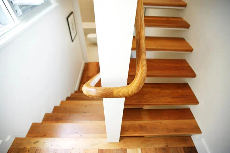 Wooden staircase with a curved wooden handrail and white wall panels, viewed from the top looking down.