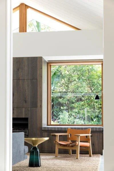 Interior of a modern living space with large window showing greenery outside, wooden accents, a small wooden bench, a black pendant light, and a decorative vase.