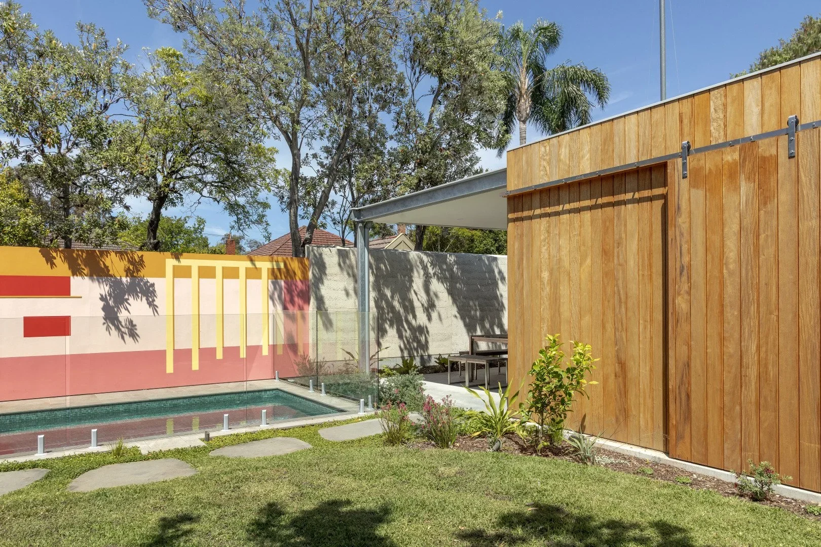 A modern backyard with a small swimming pool, a colorful mural on a stucco wall, a wooden fence, a grassy lawn, and a seating area with trees and clear blue sky overhead.