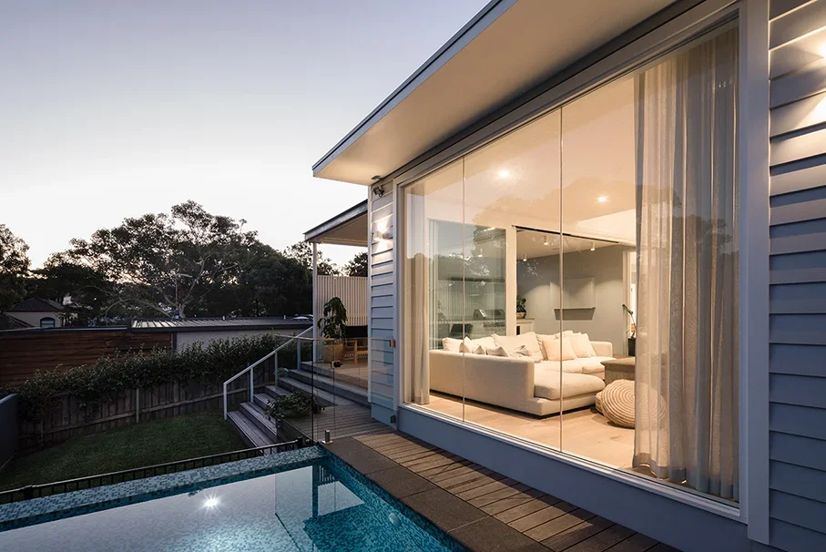 Modern house with large glass windows showing a cozy living room inside, backyard with a swimming pool, and sunset sky.