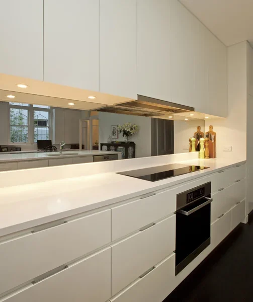 Modern white kitchen with sleek cabinetry, a built-in oven, and decorative vases on the counter, with a view of a dining area through a pass-through window.