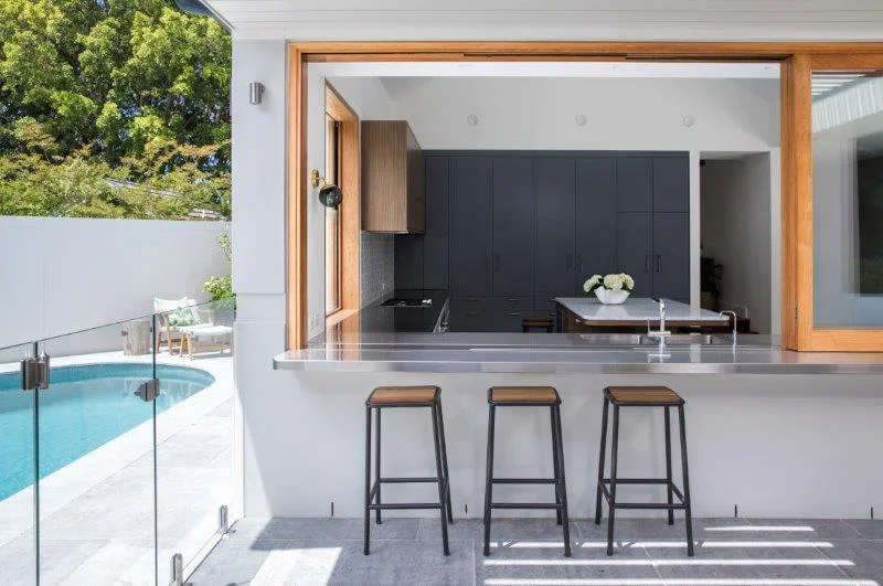 Modern kitchen with a stainless steel countertop, black cabinets, and a breakfast bar with three stools, overlooking a backyard with a swimming pool and outdoor seating.