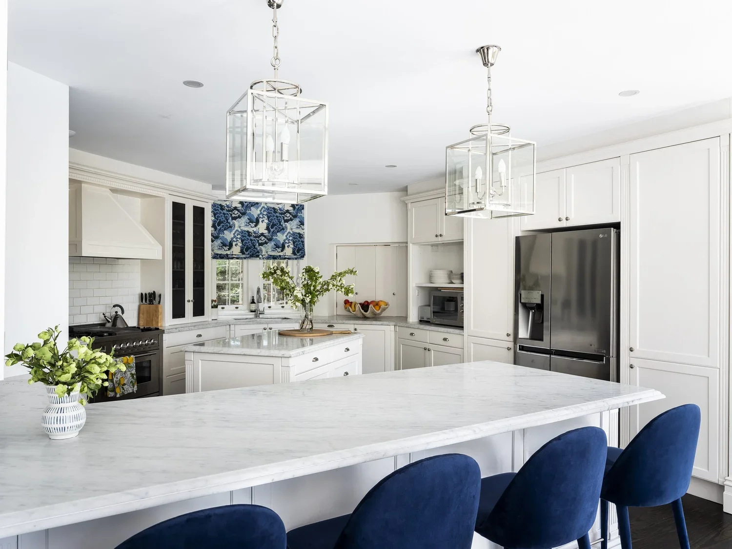 A bright, all-white kitchen with marble countertops, blue chairs at a large island, and modern pendant lights. There is a window with a blue patterned shade, a stainless steel refrigerator, and white cabinetry.