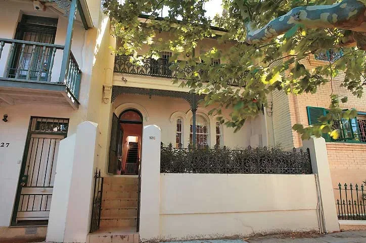 Front view of a two-story house with steps leading to an arched doorway, surrounded by a white wall and decorative black iron fence. Green foliage from a nearby tree partially covers the house.
