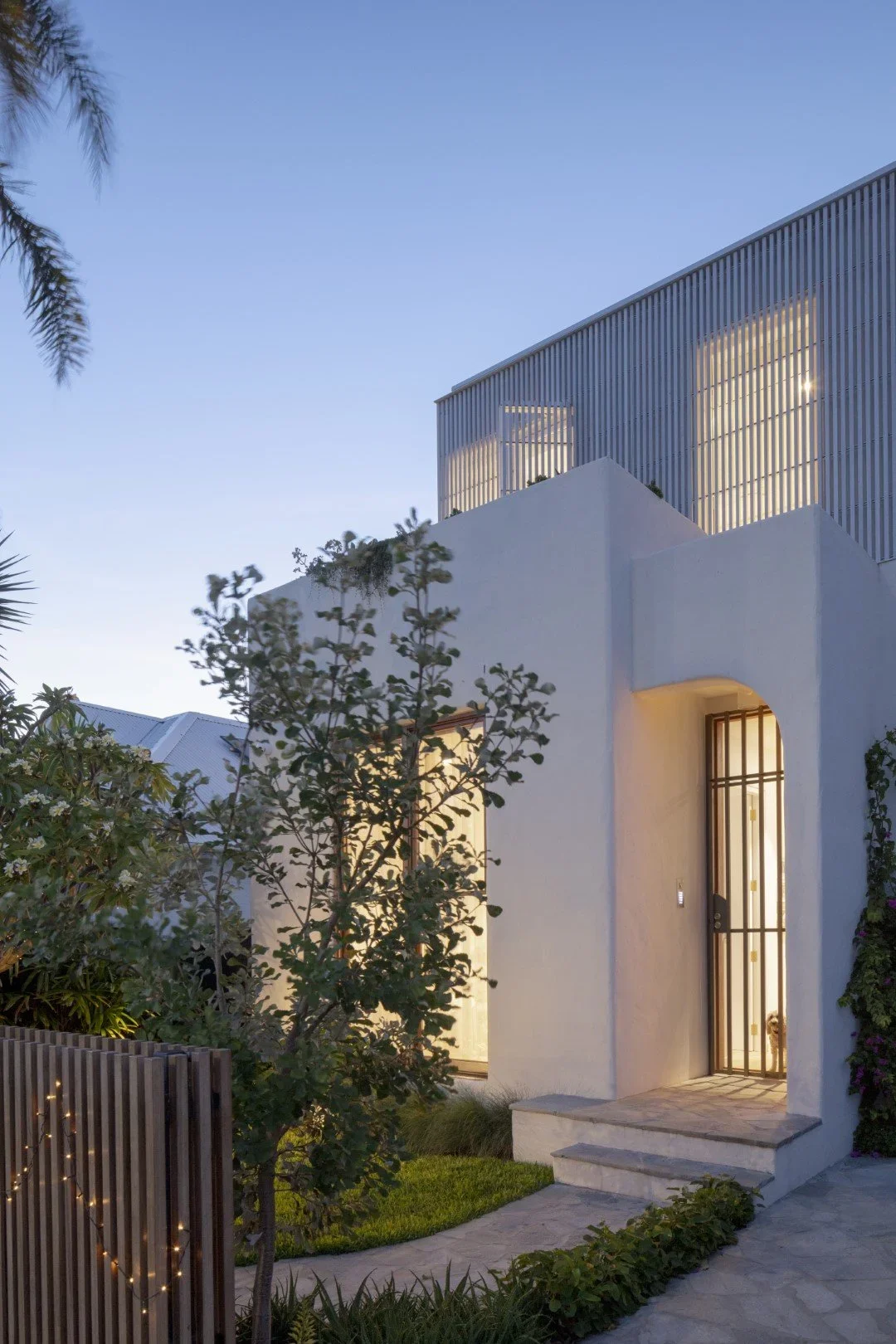 Modern white house with arched entryway, wooden door with bars, and surrounding greenery at dusk.