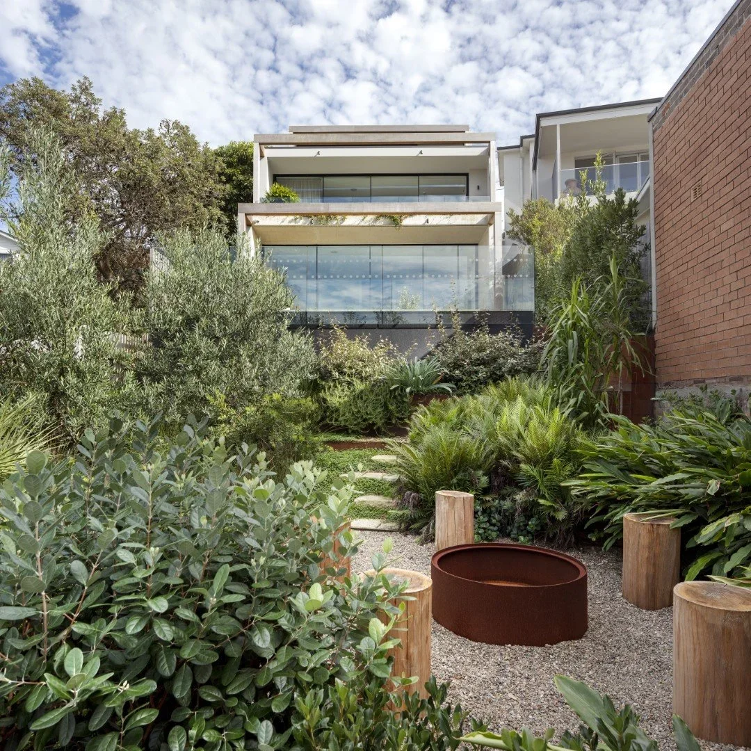 Modern multi-story house with large glass balconies, surrounded by lush green garden and stepping stones, under partly cloudy sky.