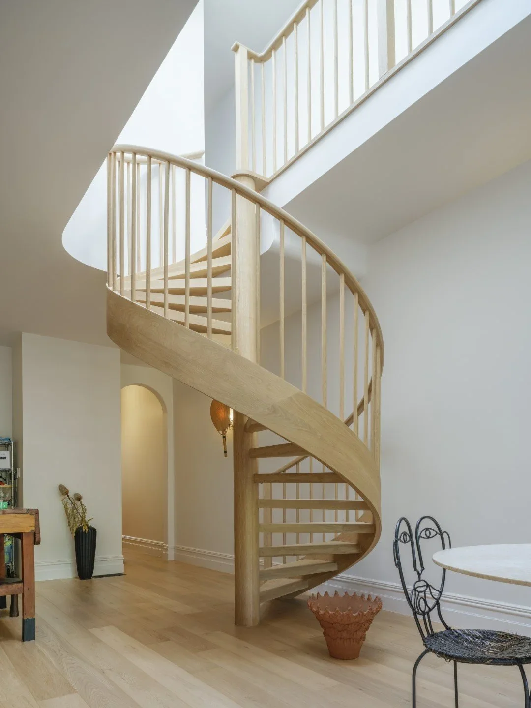 Wooden spiral staircase inside a home with light-colored walls and hardwood flooring, next to a part of a black metal chair and a round table.