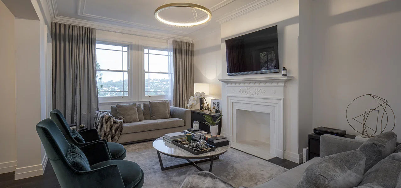 Living room with beige sofa, dark green armchairs, white fireplace, wall-mounted TV, large windows with beige curtains, and modern decor.