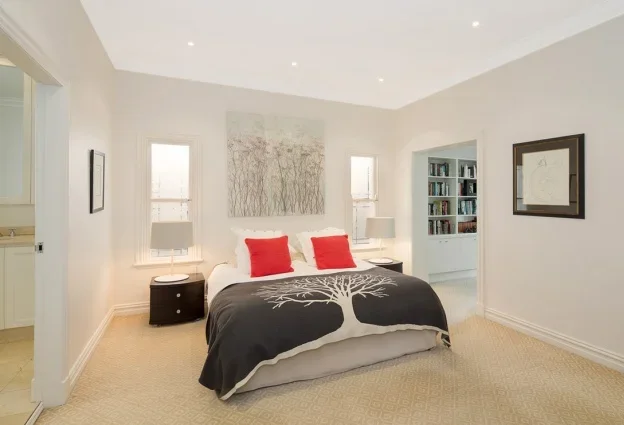 A bedroom with a bed featuring a black blanket with a white tree design, two red pillows, and two gray table lamps on bedside tables. There are two windows behind the bed, a painting above, and a bookshelf in the adjacent room.