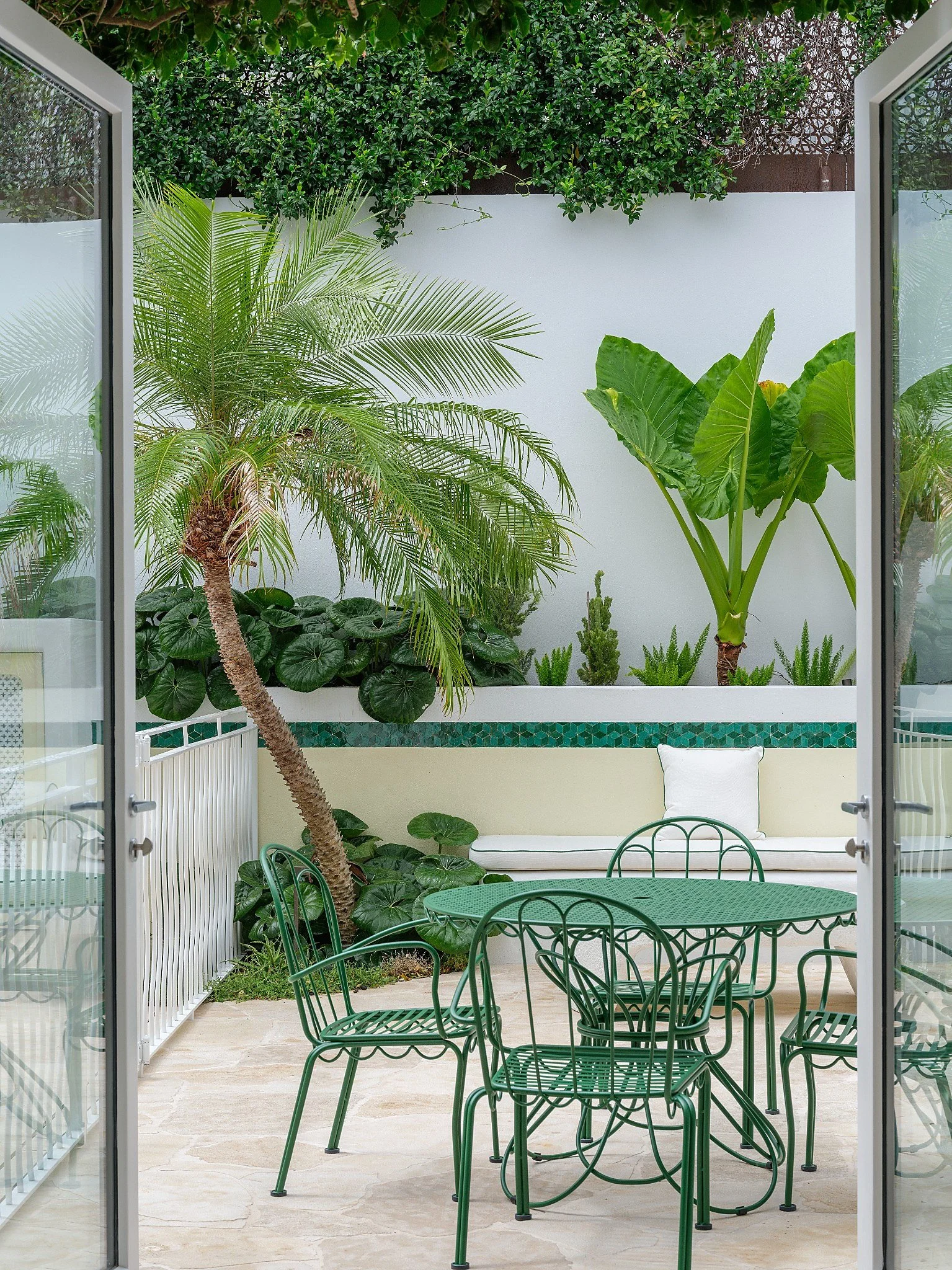 Outdoor patio with white furniture, potted plants including large green leaves, a round white coffee table, and a white wall with a sliding glass door.