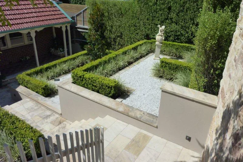 A modern, landscaped backyard with stone steps leading down to a gravel area, bordered by trimmed hedges and plants, featuring a classical statue at the far end, and partially visible house with a red-tiled roof on the left.