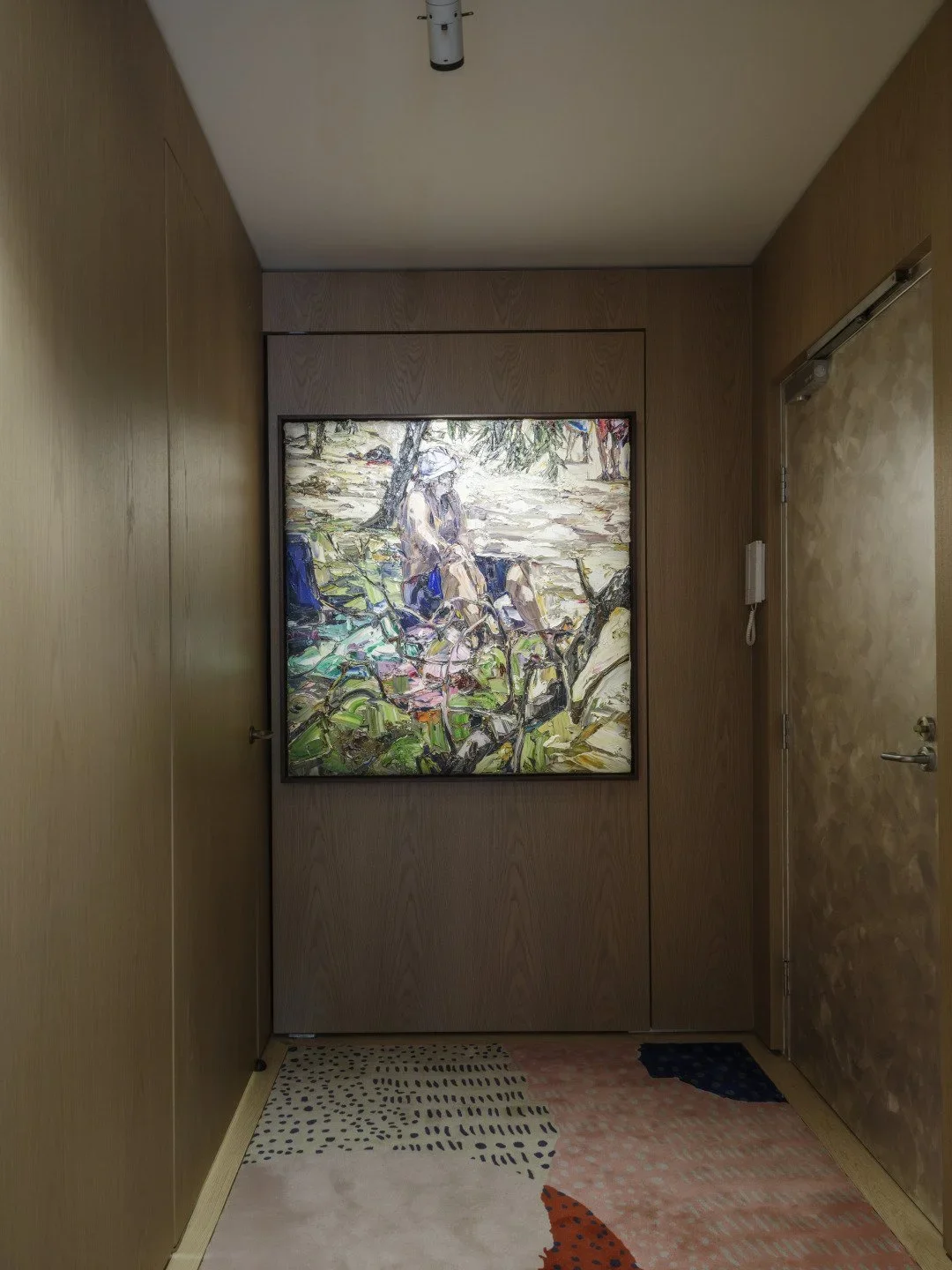 A watercolor painting of a woman sitting on a bench outdoors, surrounded by greenery and trees, displayed on a wall in a room with wooden walls and a multicolored patterned rug.