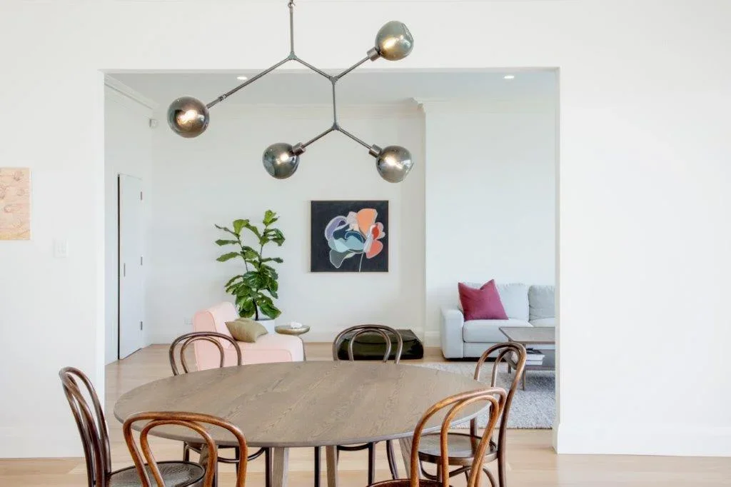 Dining area with a round wooden table surrounded by six chairs, modern chandelier overhead, sitting room with pink and gray sofas, abstract wall art, large green plant, and hardwood floors.