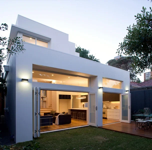 Modern white two-story house with large glass sliding doors open to a backyard patio and living space, evening lighting, trees surrounding the house.