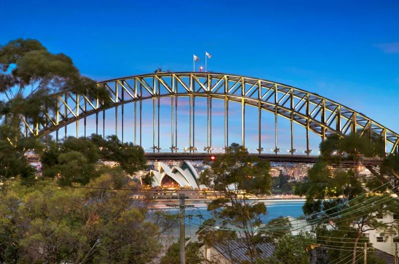 Sydney Harbour Bridge with the Sydney Opera House visible beneath it, framed by trees and power lines during dusk.