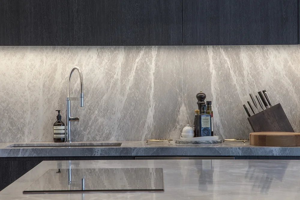 Modern kitchen countertop with a sink, soap dispenser, bottles of oil or vinegar, a knife block with knives, and a cutting board.