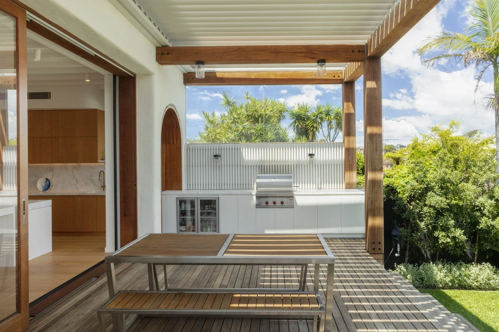 Outdoor patio with wooden deck, a built-in grill, and lush green trees in the background under a partly cloudy sky.