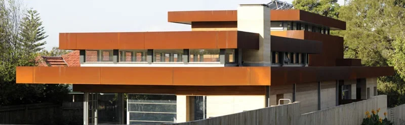 A modern multi-story house with a rust-colored exterior, flat roof, and large windows, surrounded by trees and greenery.