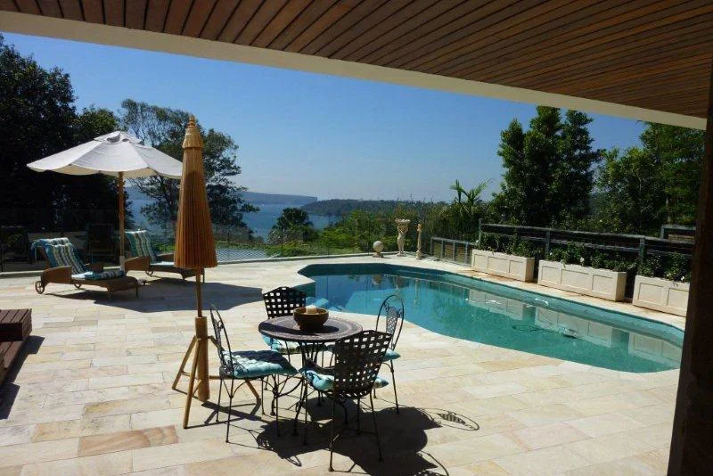 Residential backyard with a swimming pool, outdoor furniture including chairs and tables, an umbrella, and greenery with trees and a view of water and hills in the distance.