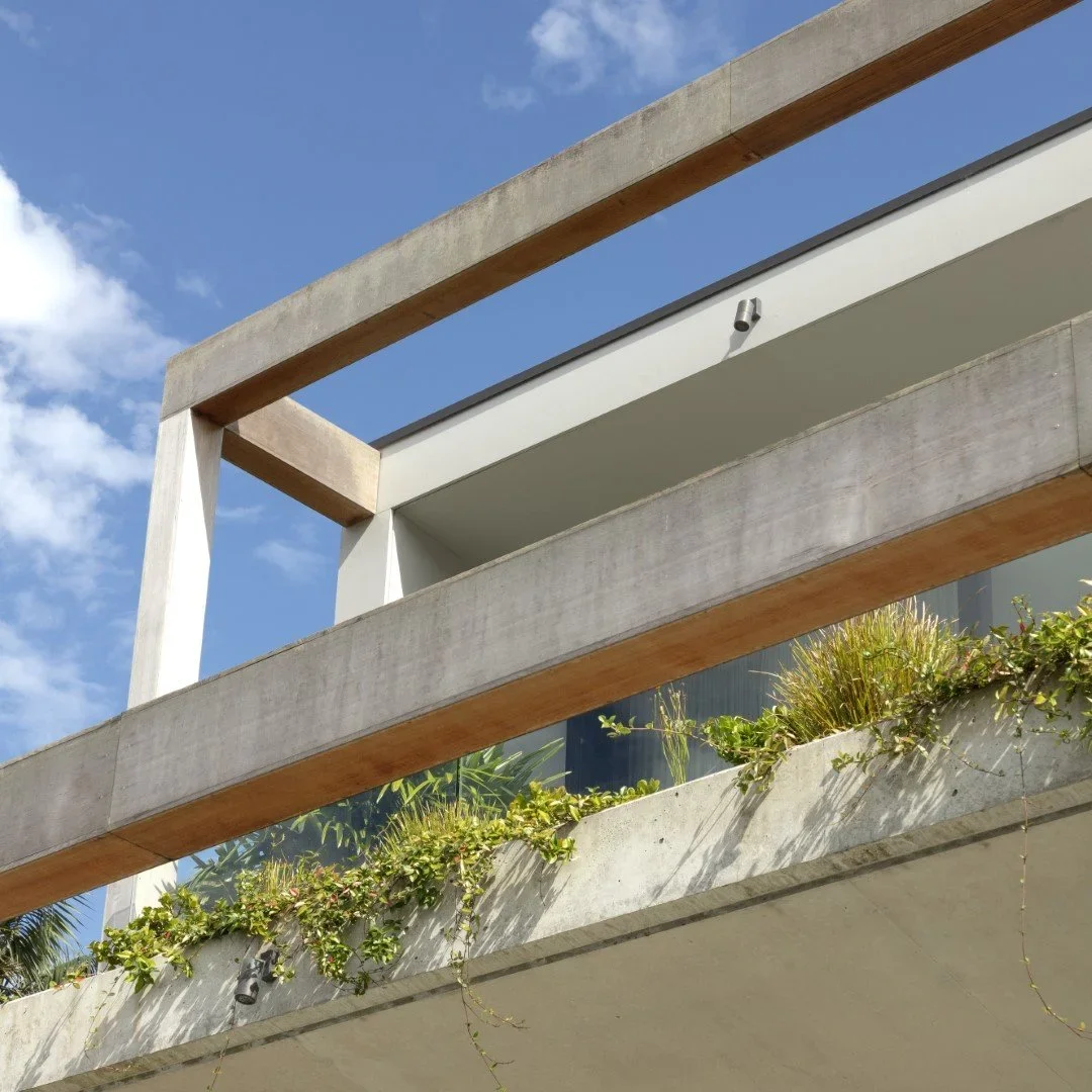 Close-up view of a modern building exterior with concrete and wood beams, greenery on the balcony, and a blue sky with some clouds in the background.