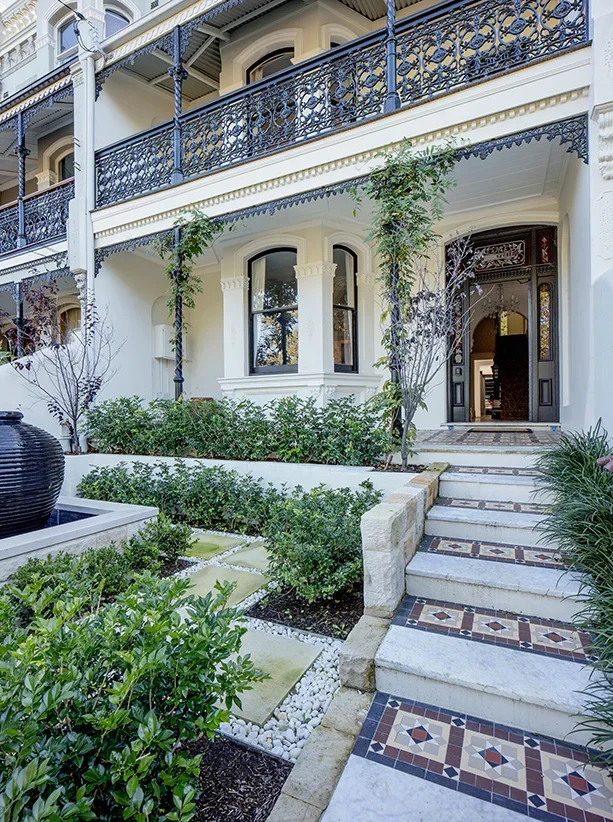 Exterior of a large, elegant house with a tiled staircase leading to the front door, surrounded by landscaped bushes, small trees, and decorative gravel.