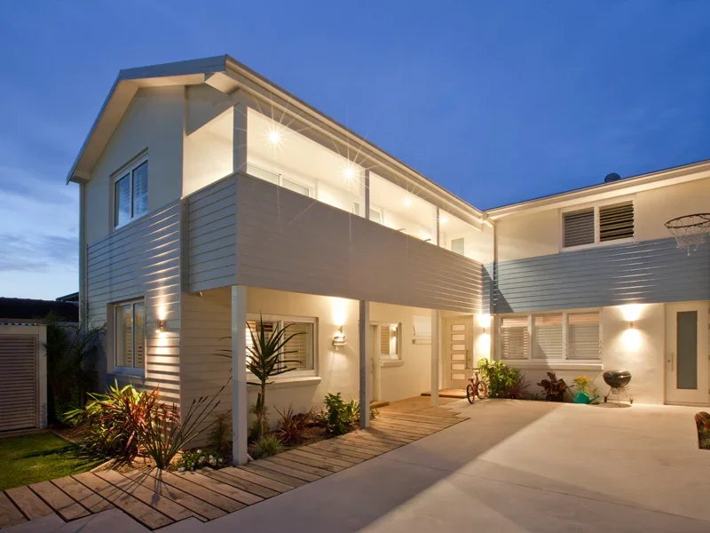 Modern two-story white house with a balcony, exterior lighting, and a small garden with plants, set against a dusk sky.