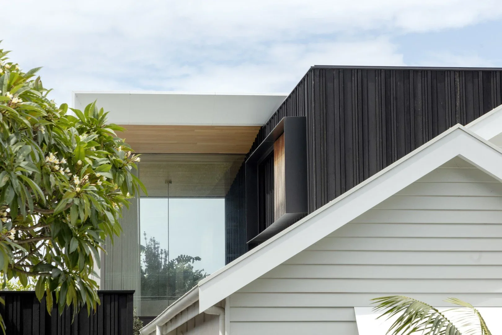 Close-up of modern house exterior with black and white siding, large window, and greenery, under a cloudy sky.