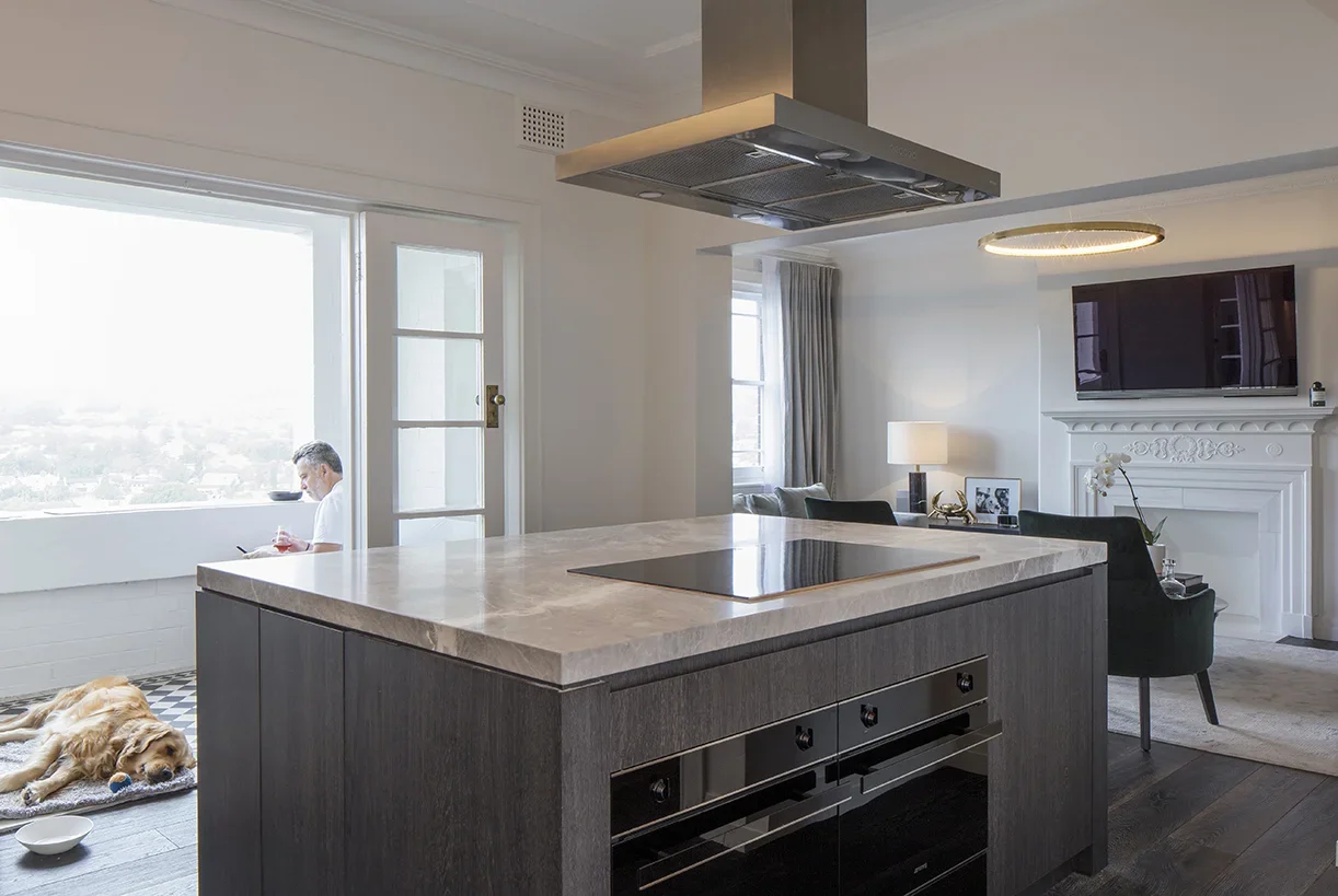 Modern open-concept living room and kitchen with large window, man sitting on window ledge, dog lying on floor, kitchen island with marble countertop, induction cooktop, and black cabinetry, sitting area with chairs, wall-mounted TV above fireplace, 