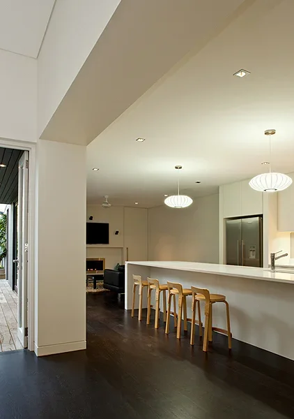 Modern open-concept kitchen and living area with white walls, island with four wooden stools, pendant lighting, and stainless steel appliances.